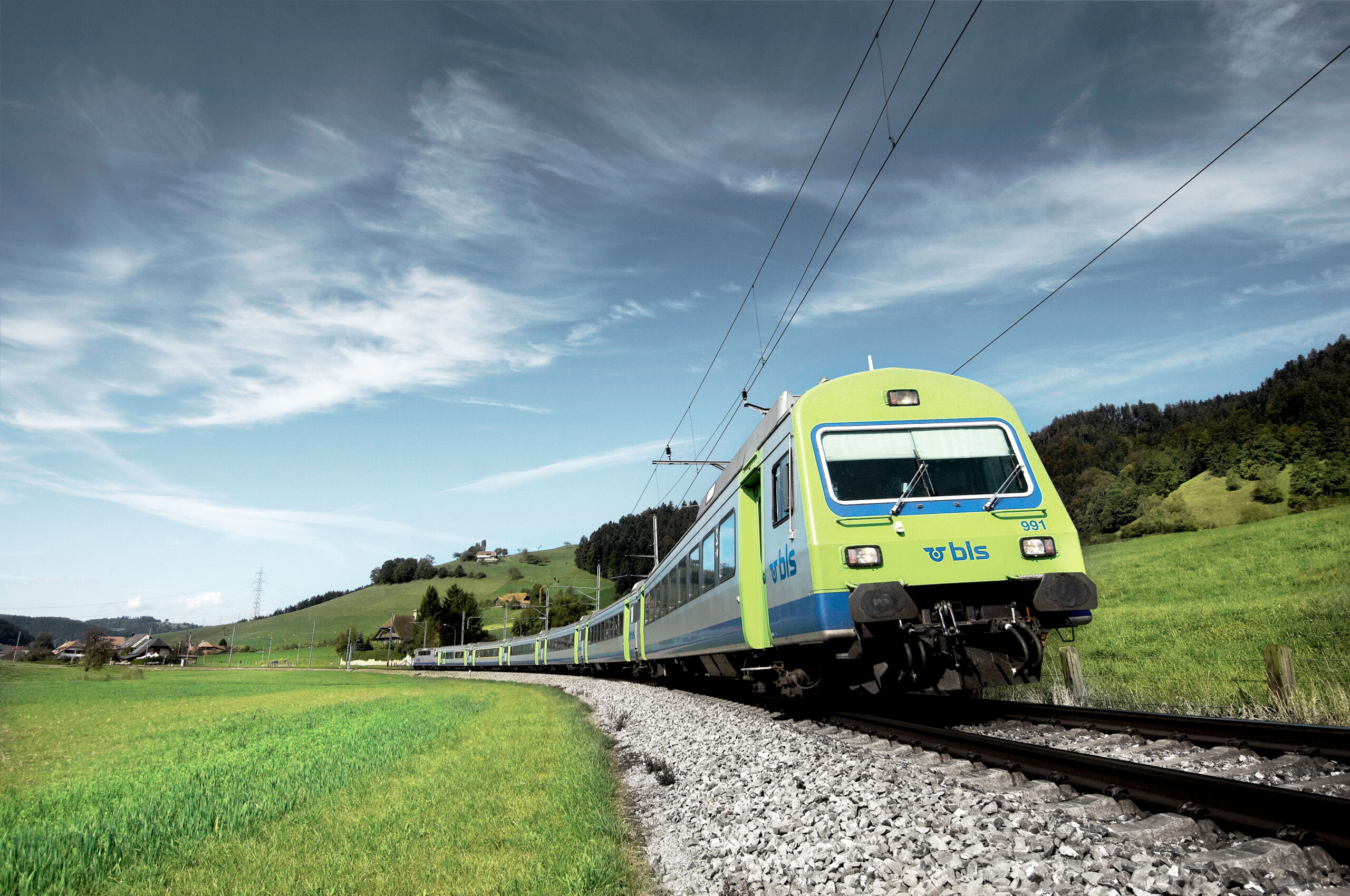 Ein grüner BLS-Zug fährt übers Land, ein paar Häuser im Hintergrund und daneben grüne Wiese