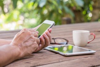 Hände tippen auf Smartphone, Tablet liegt auf Tisch neben Tasse und Brille als Symbol für BLS Case Study baseVISION